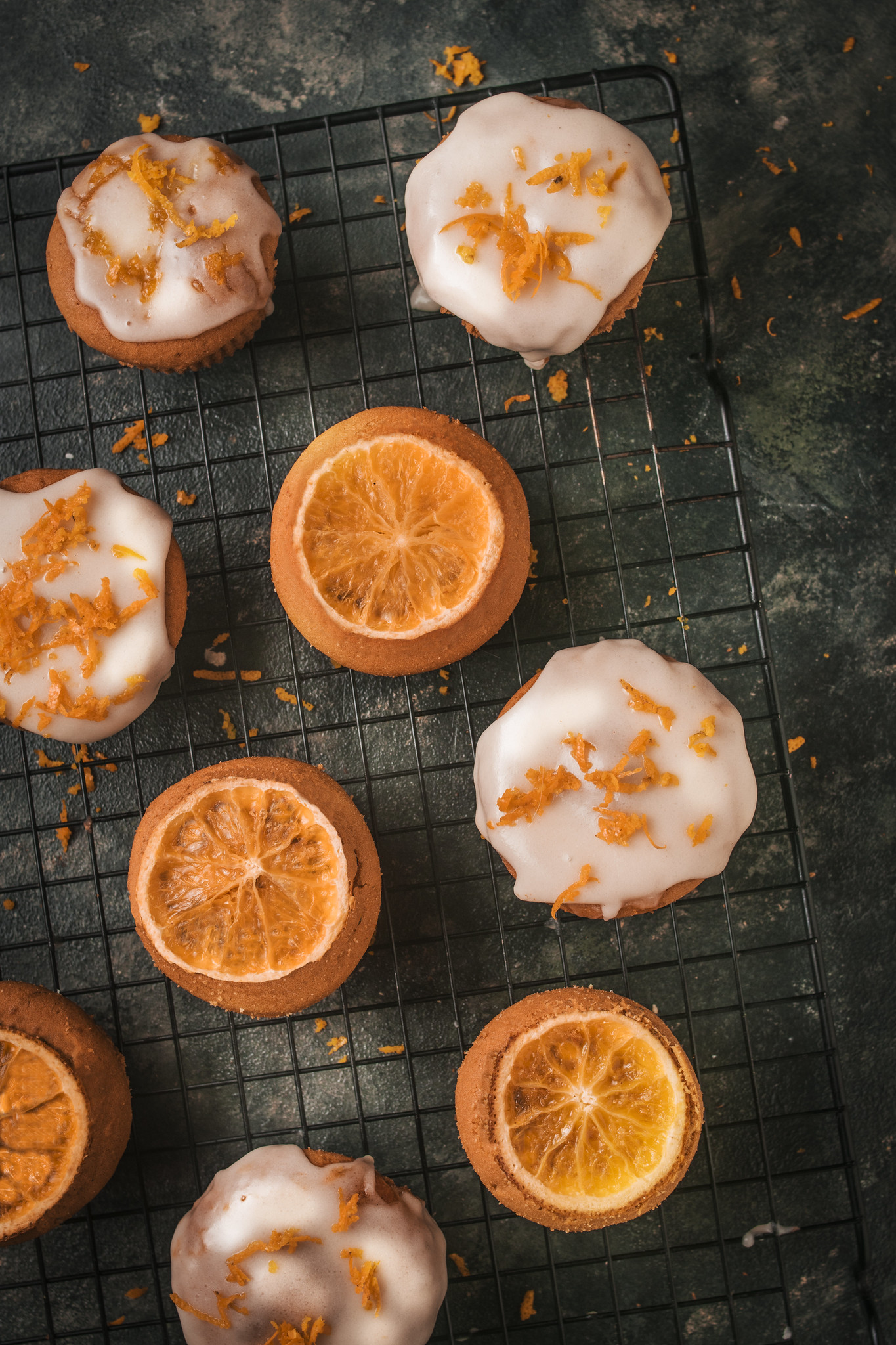 Close-up of iced blood orange cupcakes on a cooling rack, finished with a thin white glaze and scattered fresh orange zest.